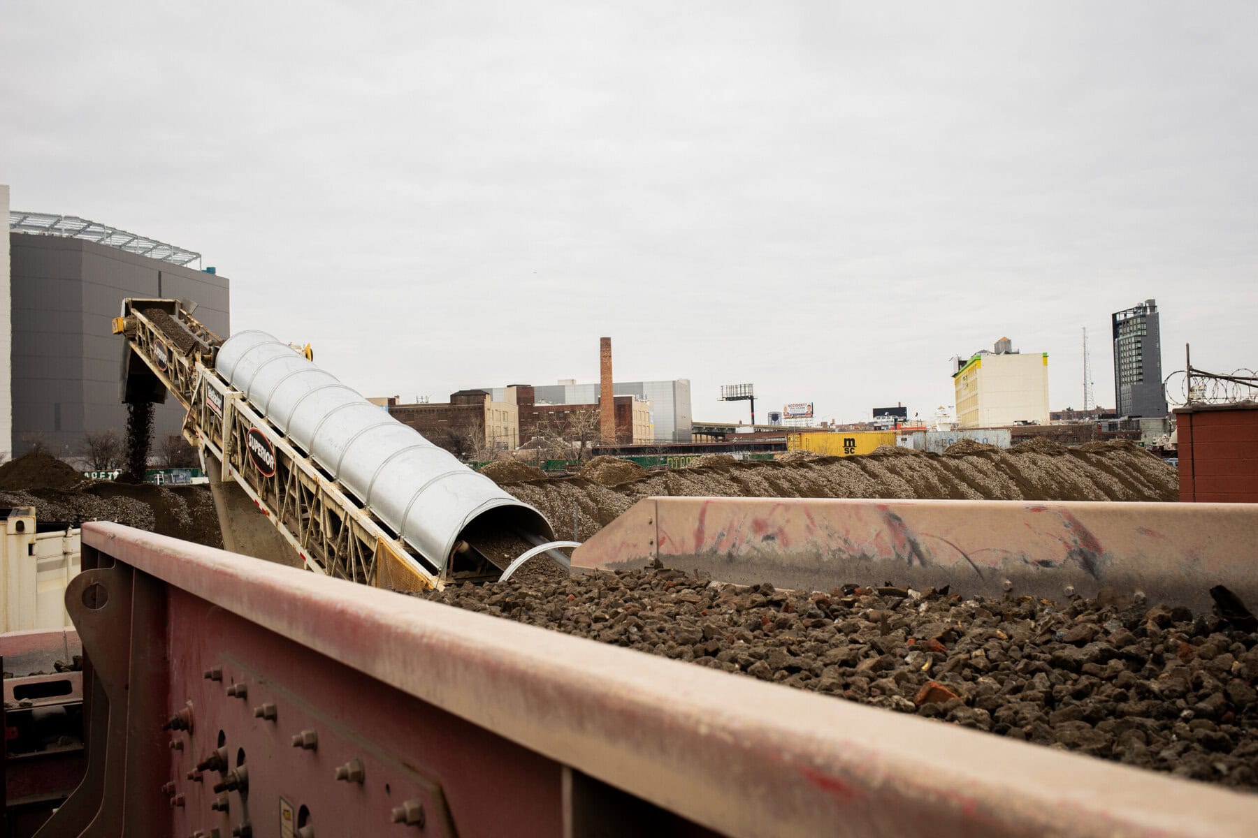 A conveyor belt moves dirt into a large container at a construction site, with city buildings and overcast sky in the background.
