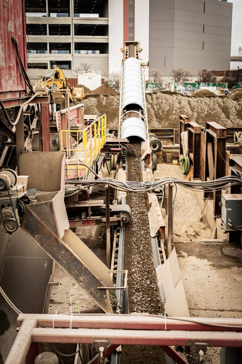 A conveyor belt transports gravel at an industrial site, surrounded by machinery and buildings in the background.