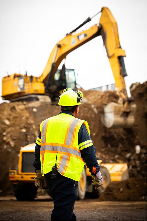 A construction worker in a high-visibility vest and helmet stands in front of a large yellow excavator at an active construction site.
