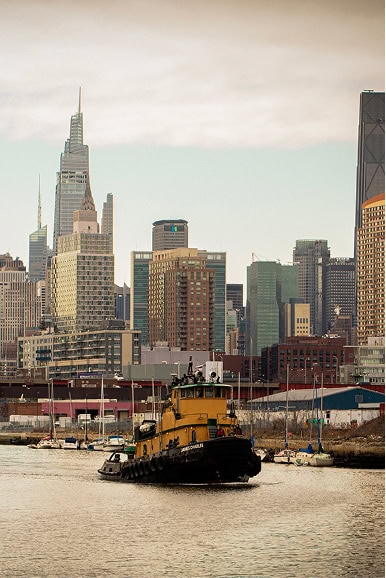 A yellow and black tugboat moves along a river with a city skyline of tall modern buildings in the background.