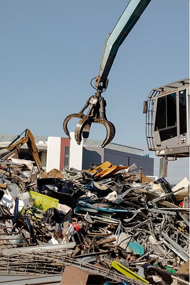 A mechanical claw attached to an excavator lifts scrap metal and debris at a junkyard, with buildings visible in the background under a clear sky.