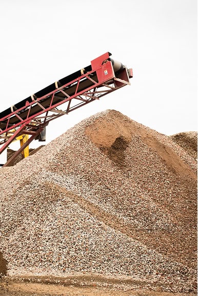 A conveyor belt deposits rocks and gravel onto a large pile at an outdoor industrial site, under an overcast sky.