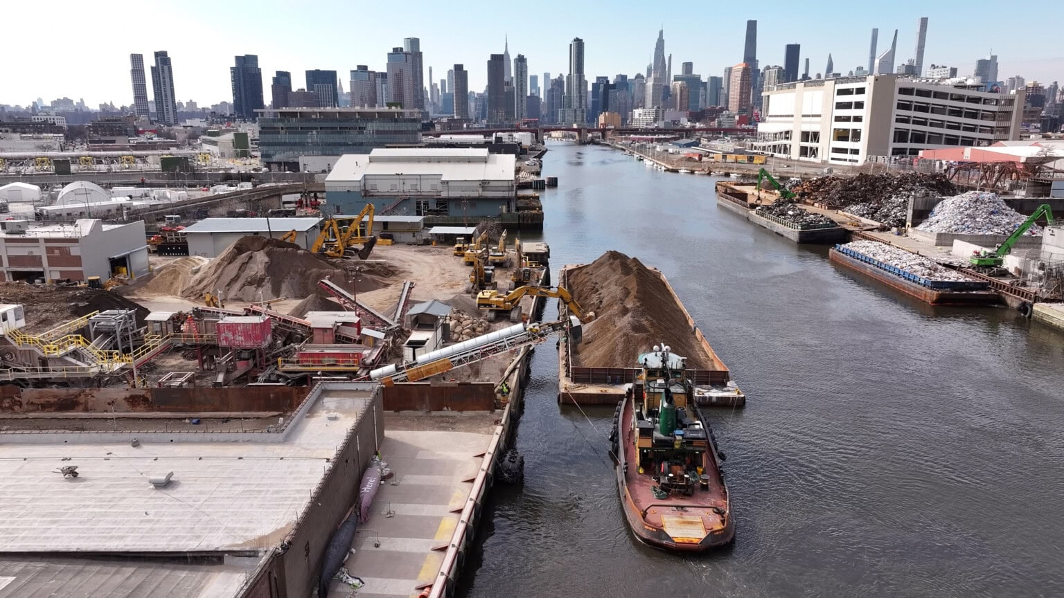 A tugboat moves a barge loaded with dirt or sand in an industrial canal, with construction equipment and piles of materials on both sides and a city skyline in the background.