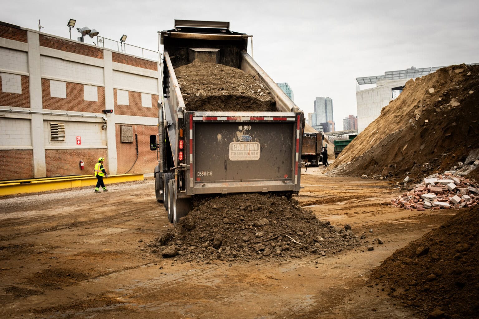 A dump truck unloads a large pile of soil at a construction site, with a worker walking nearby and city buildings in the background.
