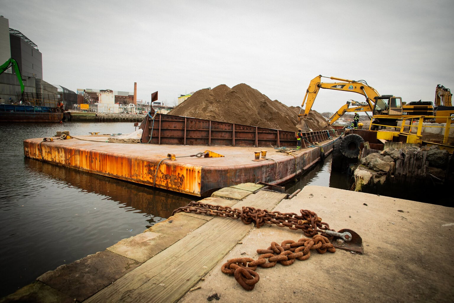 A rusted barge loaded with sand is docked at a waterfront as a yellow excavator unloads material; chains lie on the concrete pier in the foreground.