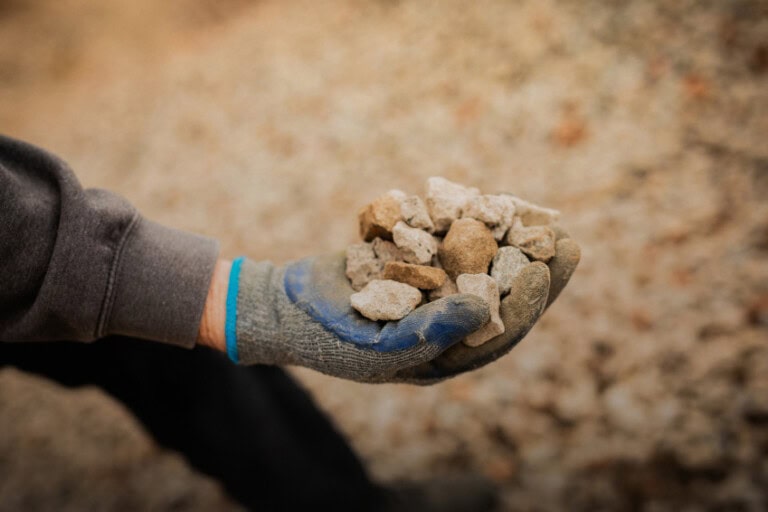 A person wearing a grey and blue work glove holds a handful of small rocks and gravel.