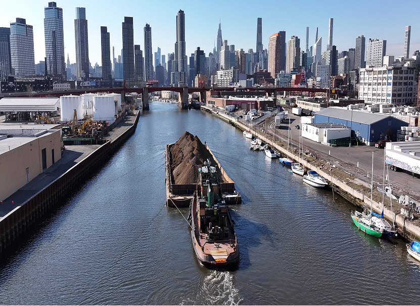 A barge carrying a load of dirt is towed along a canal, with a city skyline and numerous skyscrapers in the background. Several boats are docked along the right bank.