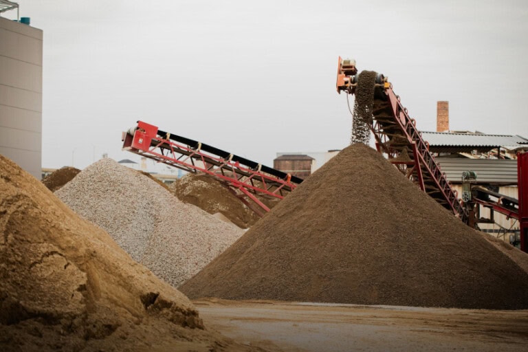 Two conveyor belts deposit gravel and sand into large piles at an industrial site, with buildings visible in the background.