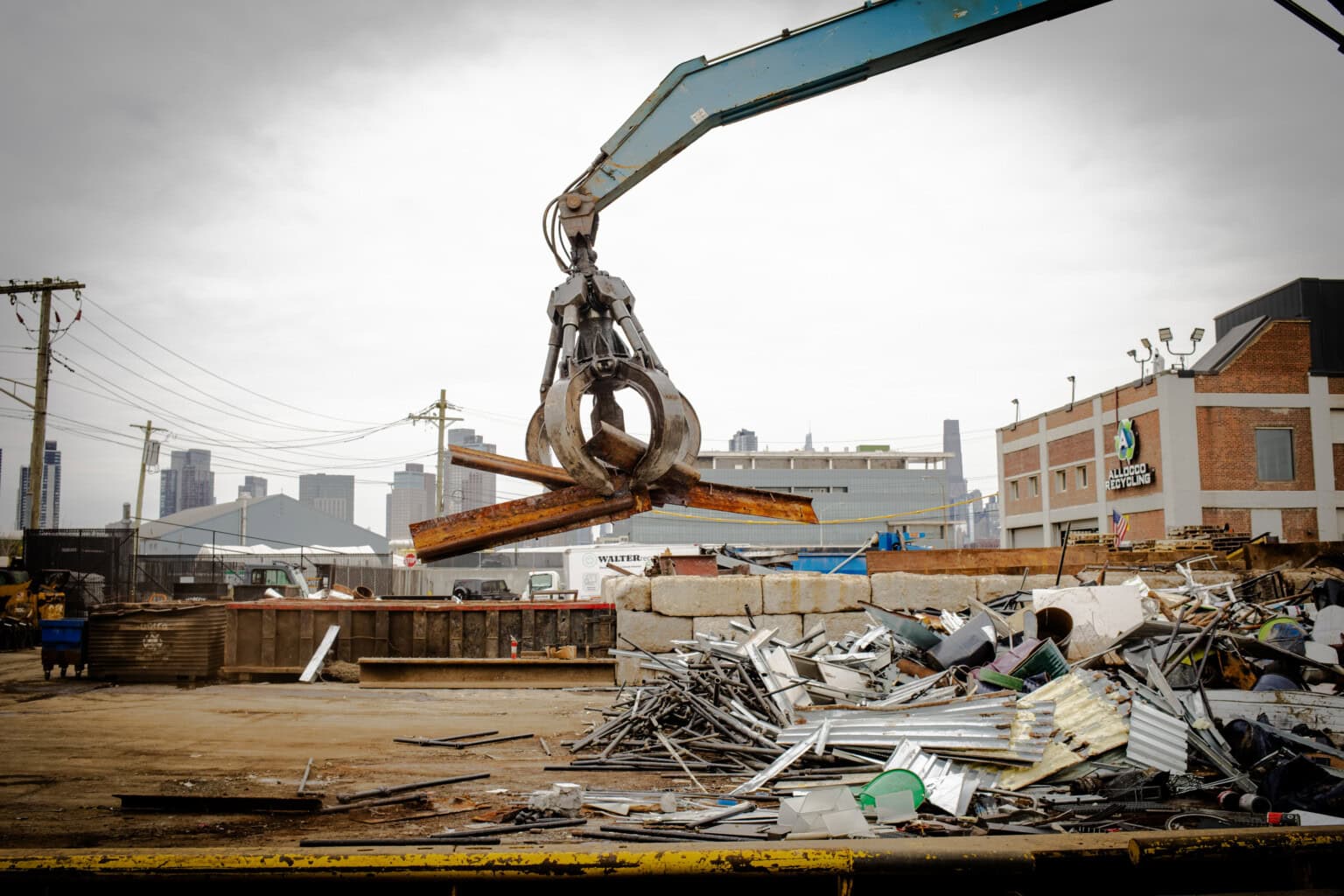 A large industrial claw lifts scrap metal at a junkyard, with piles of metal debris and buildings visible in the background under a cloudy sky.